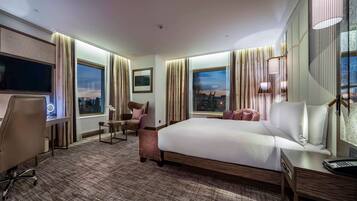 Habitación de hotel con una cama grande, un escritorio con silla, televisor y vistas a la ciudad desde las ventanas.