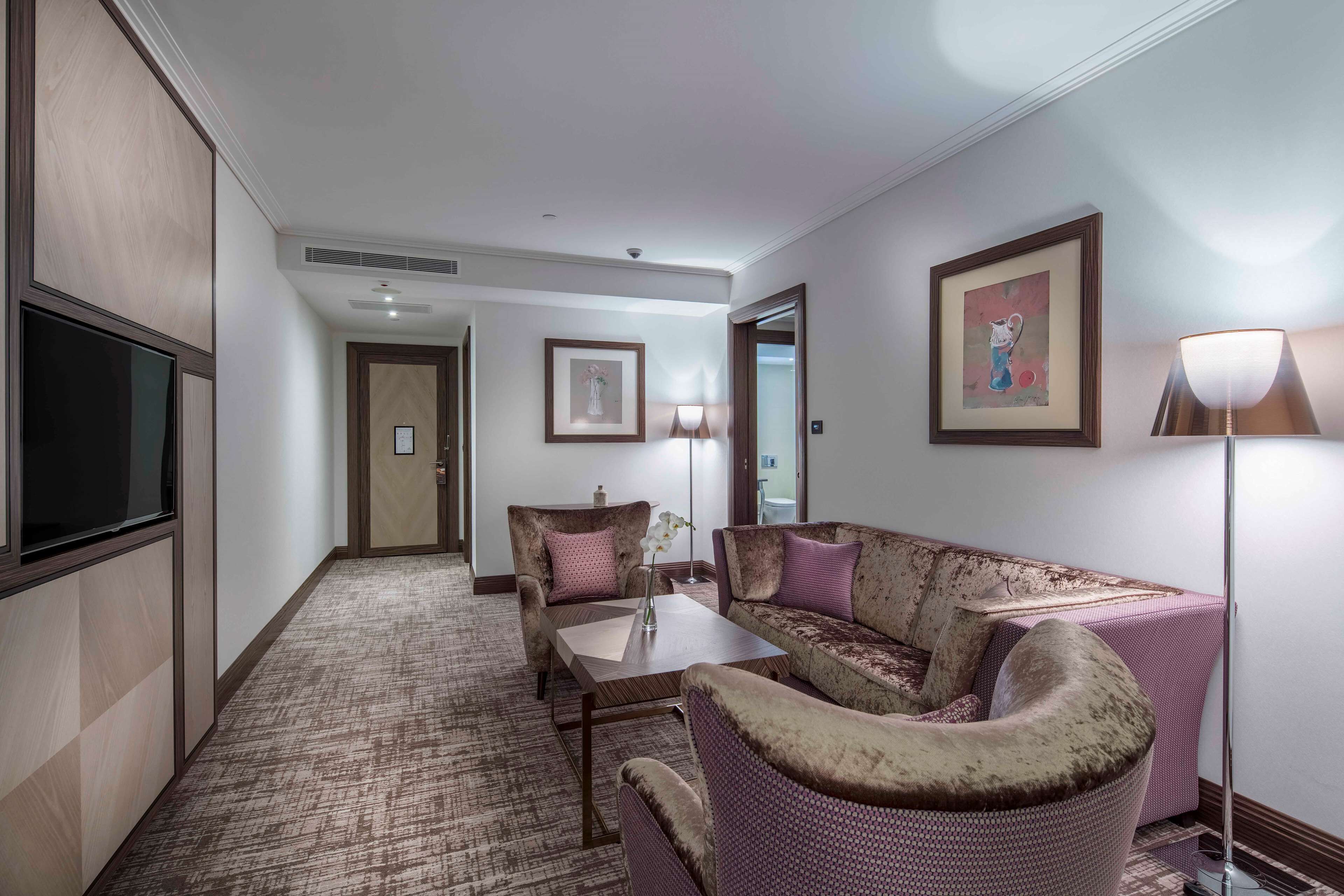 A hotel room with a seating area featuring a sofa, armchairs, and a coffee table. There is a flat-screen TV mounted on the wall, a framed picture, and a floor lamp.