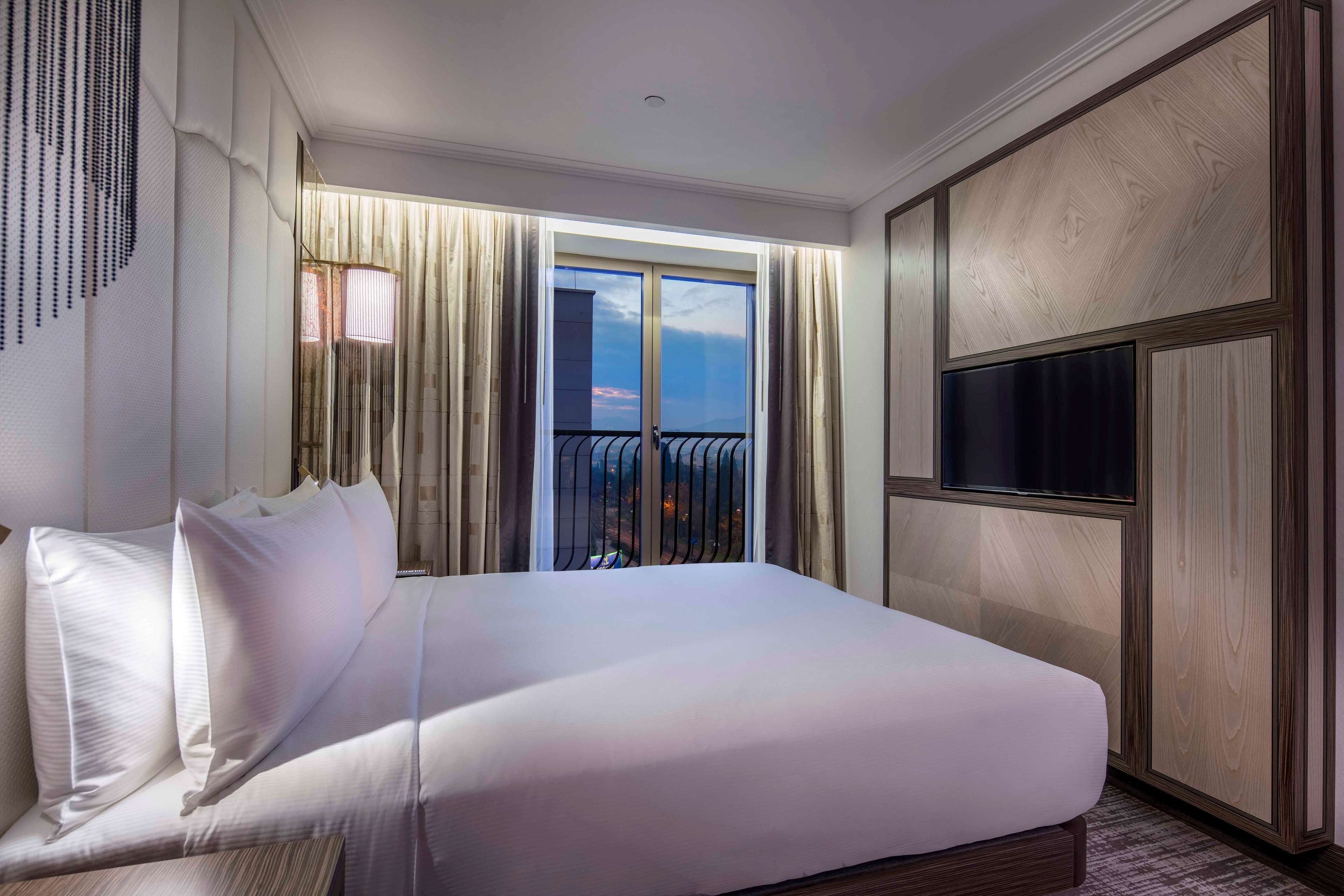 A hotel room with a large bed, a TV, a balcony with a view, and a wooden wardrobe.