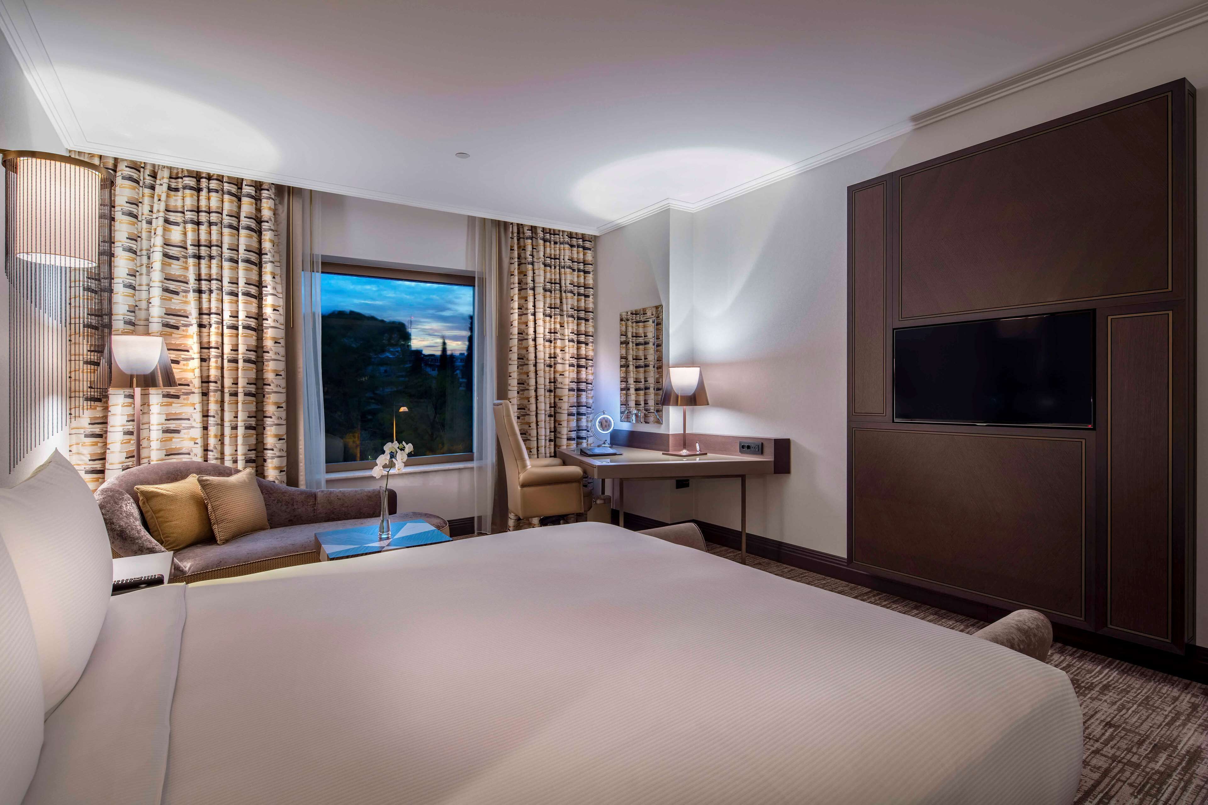 Una habitación de hotel con una cama grande, un escritorio con lámpara, una silla y un televisor montado en la pared.