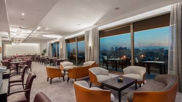 A modern hotel lobby with a city view, spacious seating, and a bar area.
