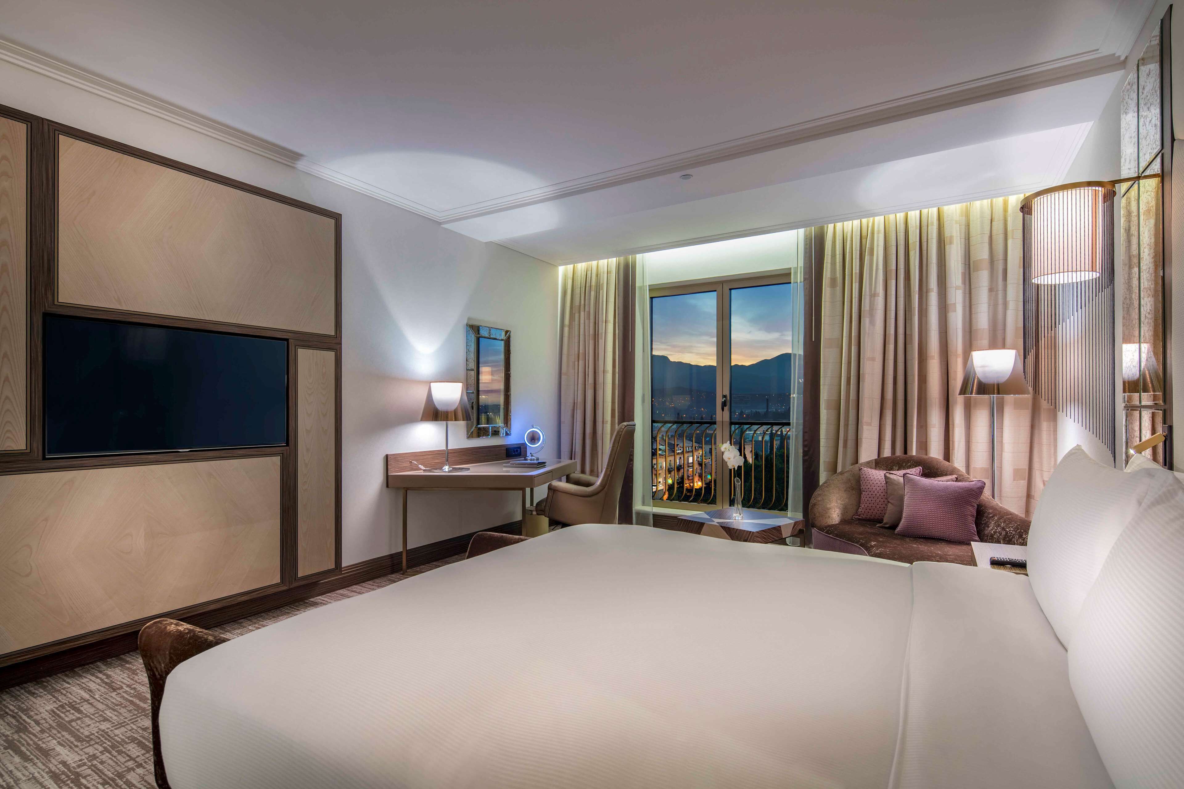 A hotel room with a large bed, a desk with a chair, a TV, a window with curtains, and a balcony with a view.