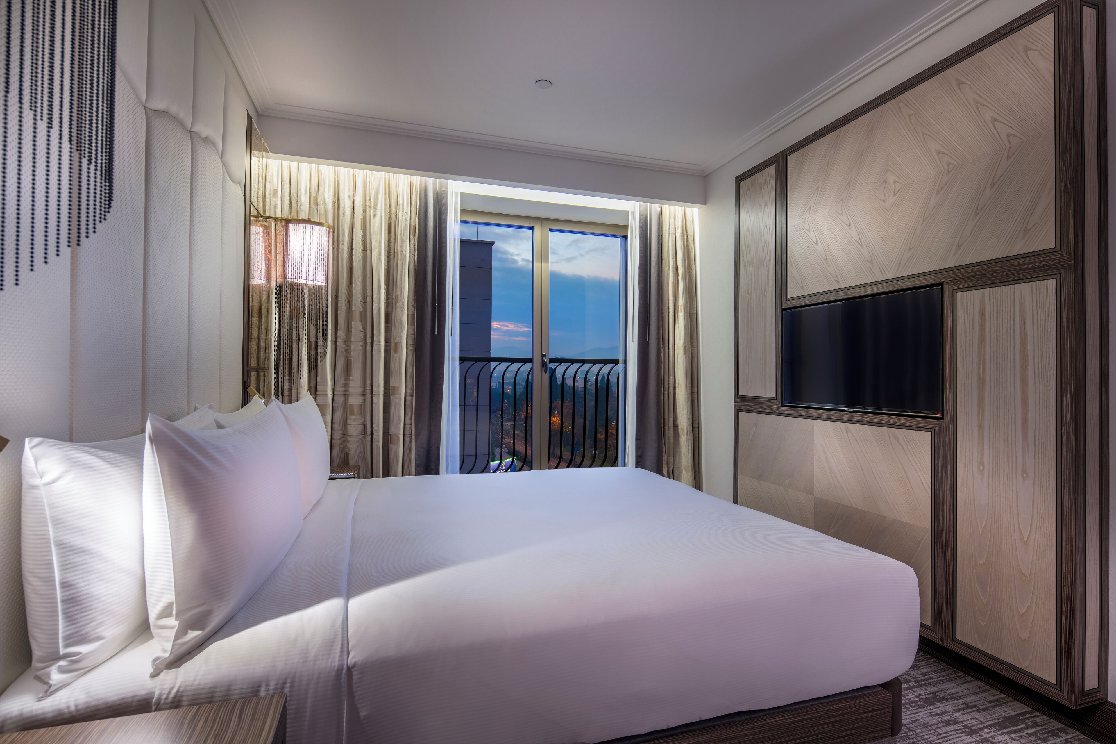 Habitación de hotel con una cama grande, televisión, balcón con barandilla y vista a la ciudad.