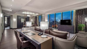 A modern hotel room with a large window offering a city view, a desk with a telephone, and a seating area with leather chairs.