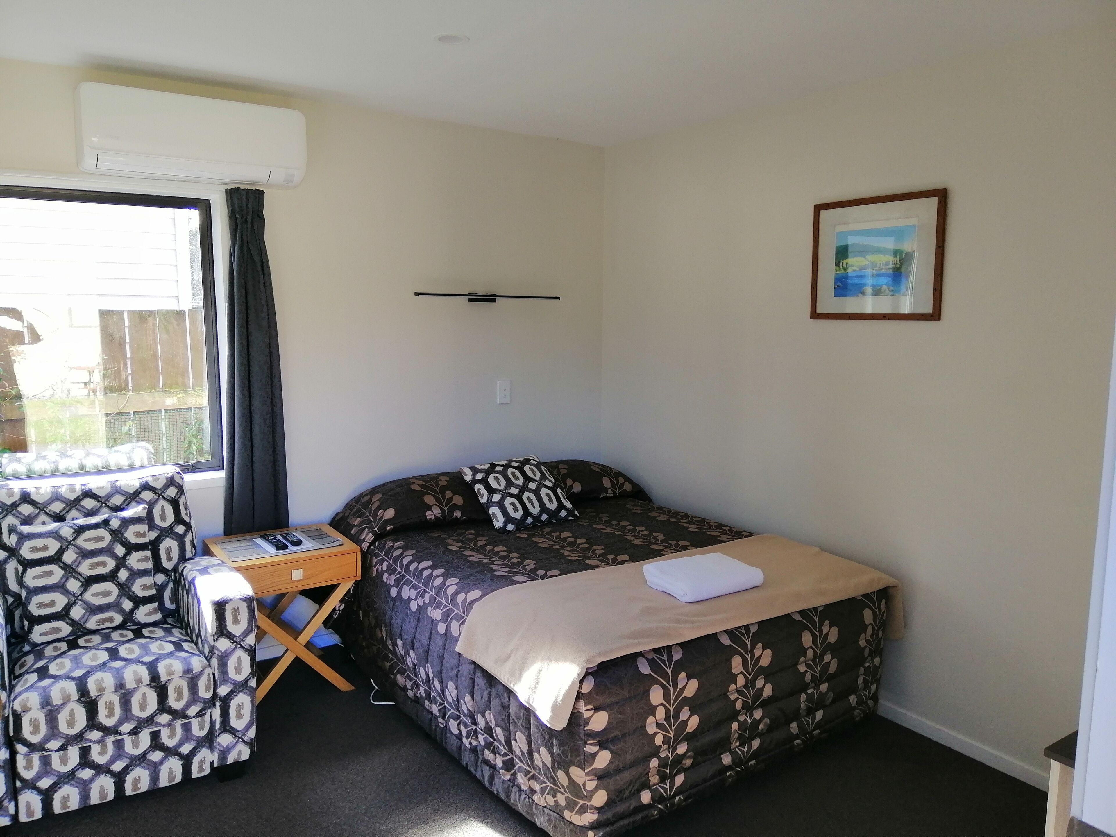 A small, well-lit room with a bed, a chair, a small table, and a window.