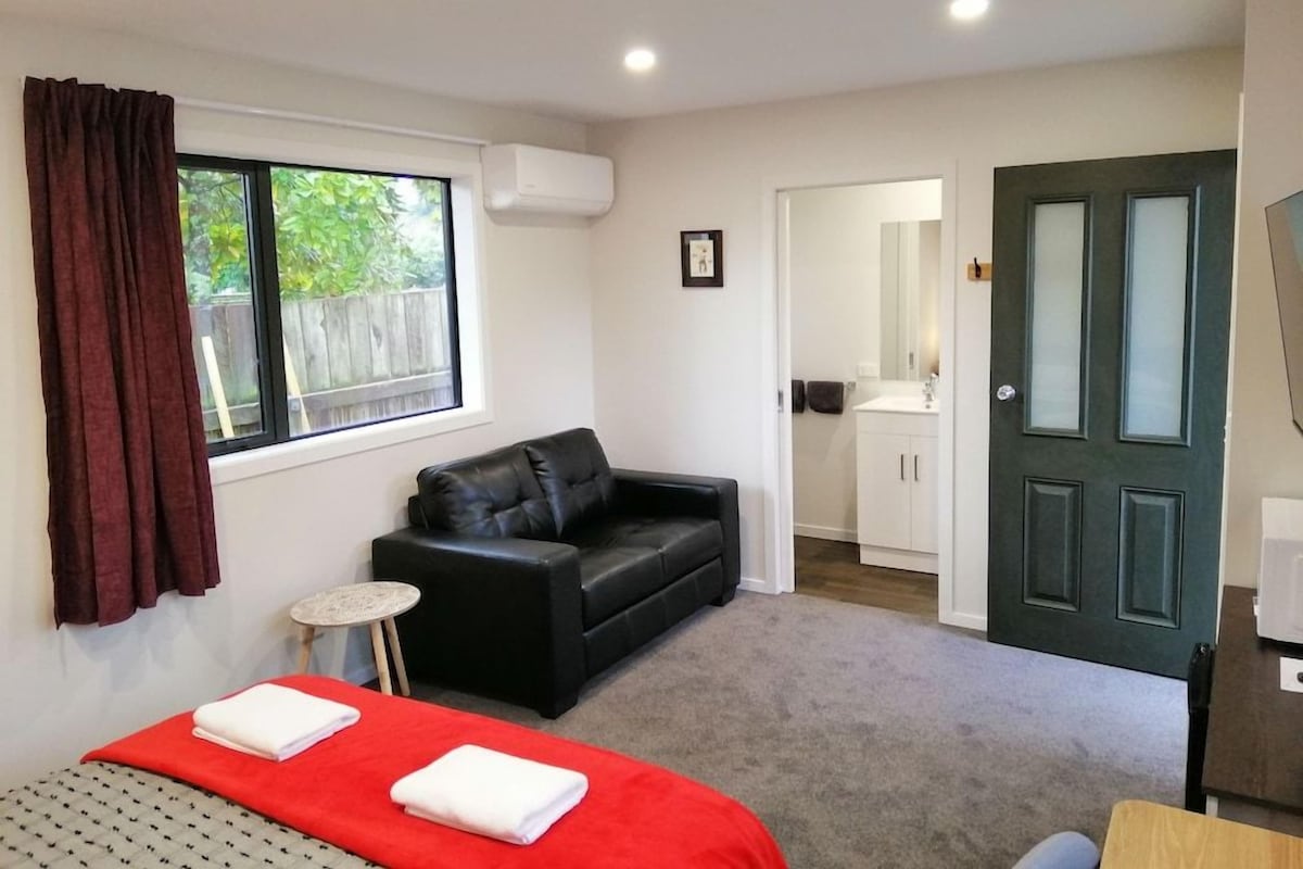 A bedroom with a bed, a black leather sofa, a small round table, and a door leading to a bathroom.