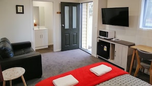 Habitación de hotel con cama roja, sofá de cuero negro, una mesita redonda pequeña, televisor montado en la pared y un área de kitchenette.