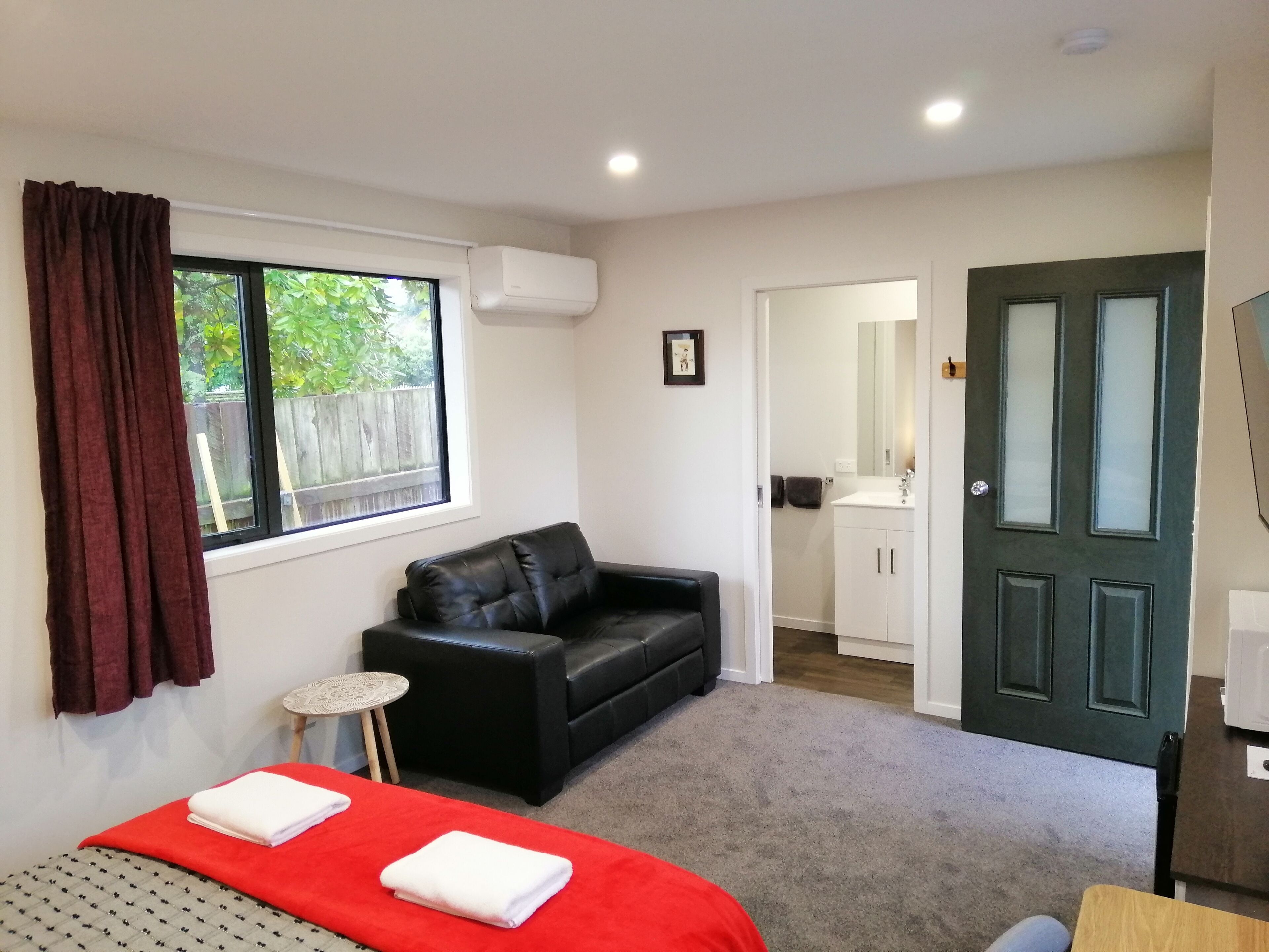 A bedroom with a bed, a black leather sofa, a small table, and a bathroom visible through an open door.