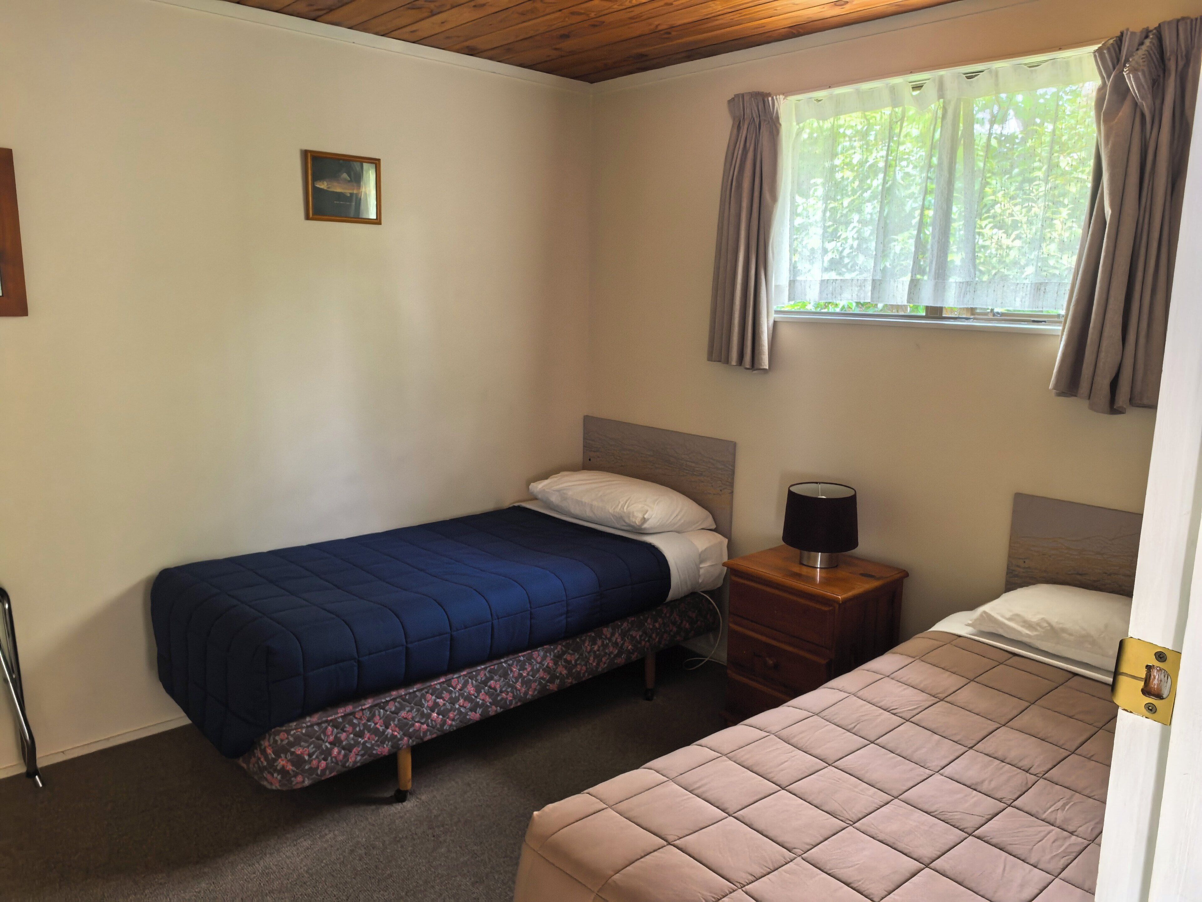 A bedroom with two beds, a window with curtains, a bedside table with a lamp, and a picture on the wall.