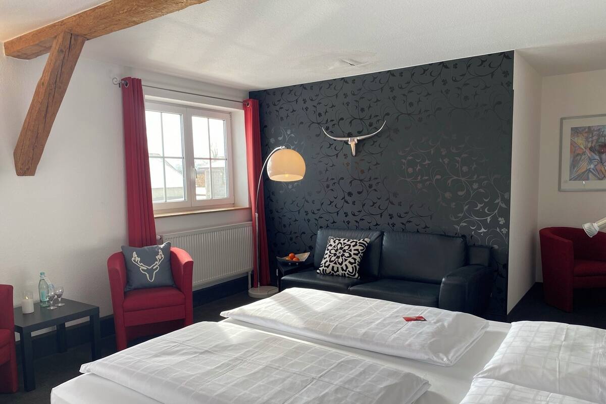 A hotel room with a large bed, a sofa, a red chair, a floor lamp, and a wall with a decorative pattern.