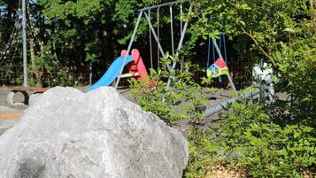 Ein Spielplatz mit Schaukeln und Rutschen, umgeben von viel Grün und einem großen Felsen.