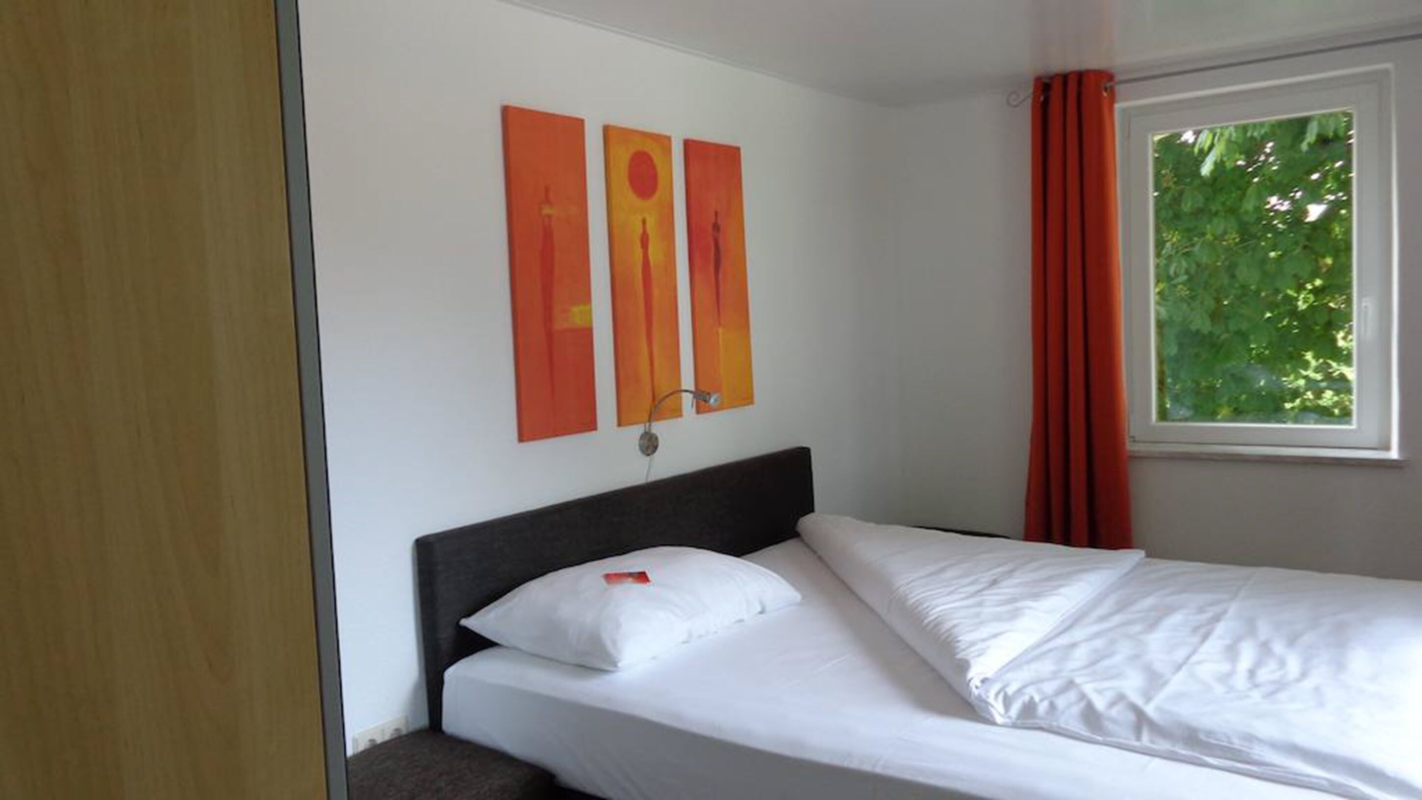 A hotel room with a single bed, white bedding, and two abstract paintings on the wall.