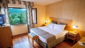 Habitación de hotel con una cama grande, mesitas de noche con lámparas, un banco de madera, un televisor y una ventana con cortinas.