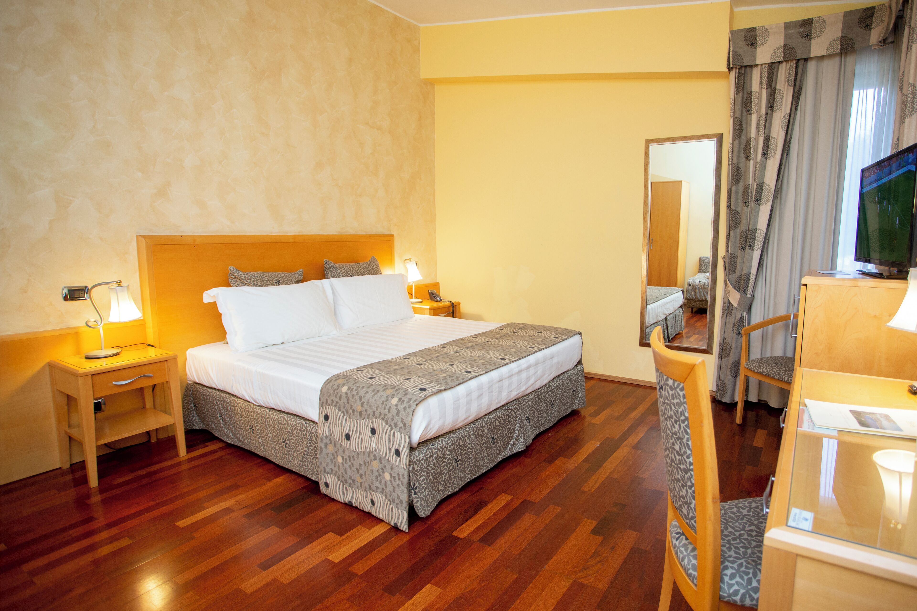 Una camera d'albergo con pavimento in legno, un letto con copripiumino fantasia, un comodino con lampada, una sedia e una televisione su un mobiletto in legno.