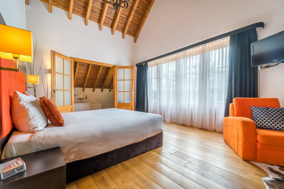 A bedroom with a bed, a TV, a chair, and a window with curtains.