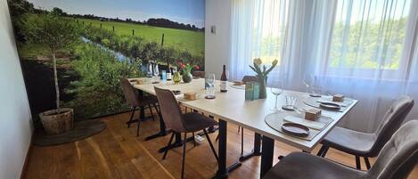 A dining area with a table set for four, chairs, a painting of a landscape, and a window with curtains.