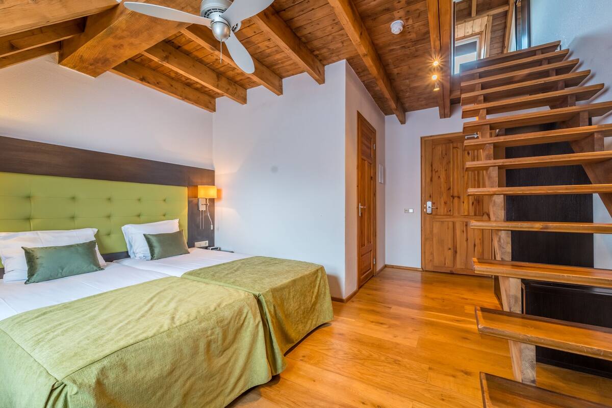 A bedroom with two beds, wooden ceiling beams, a green upholstered headboard, a wooden staircase, and a nightstand with a lamp.