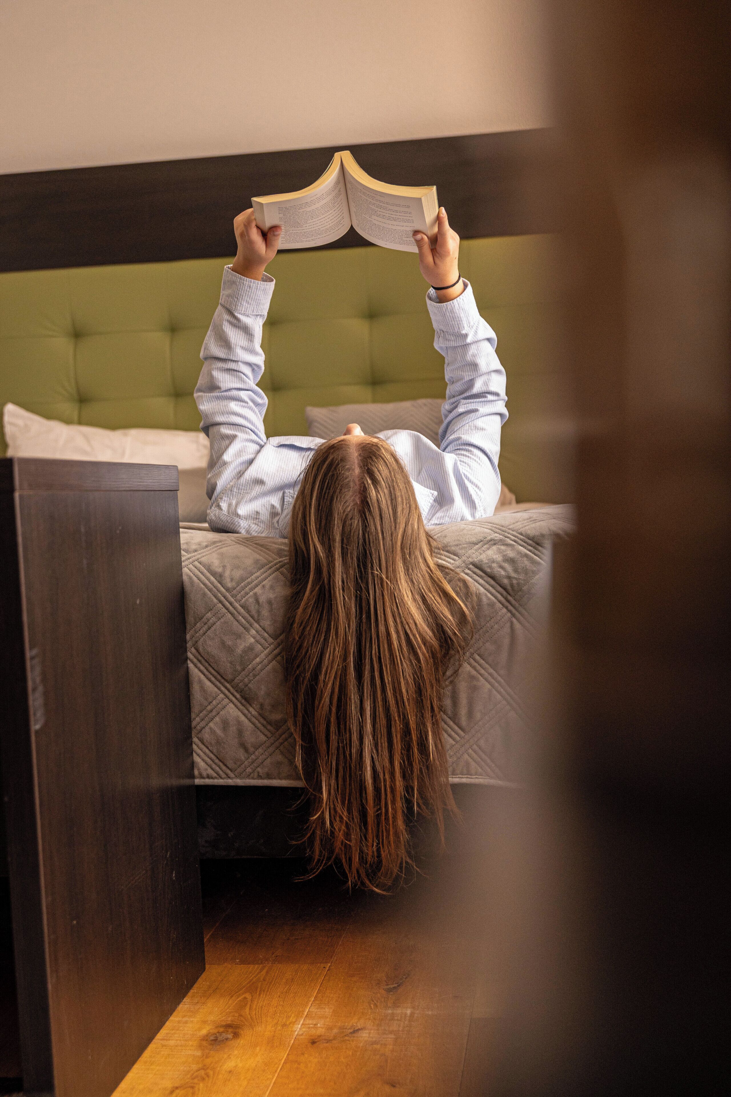 A person lying on a bed with a book, in a room with a green upholstered headboard and wooden flooring.