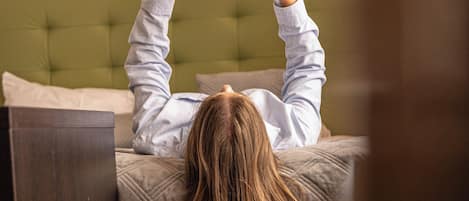 A person lying on a bed with a book, in a room with a green upholstered headboard and wooden flooring.