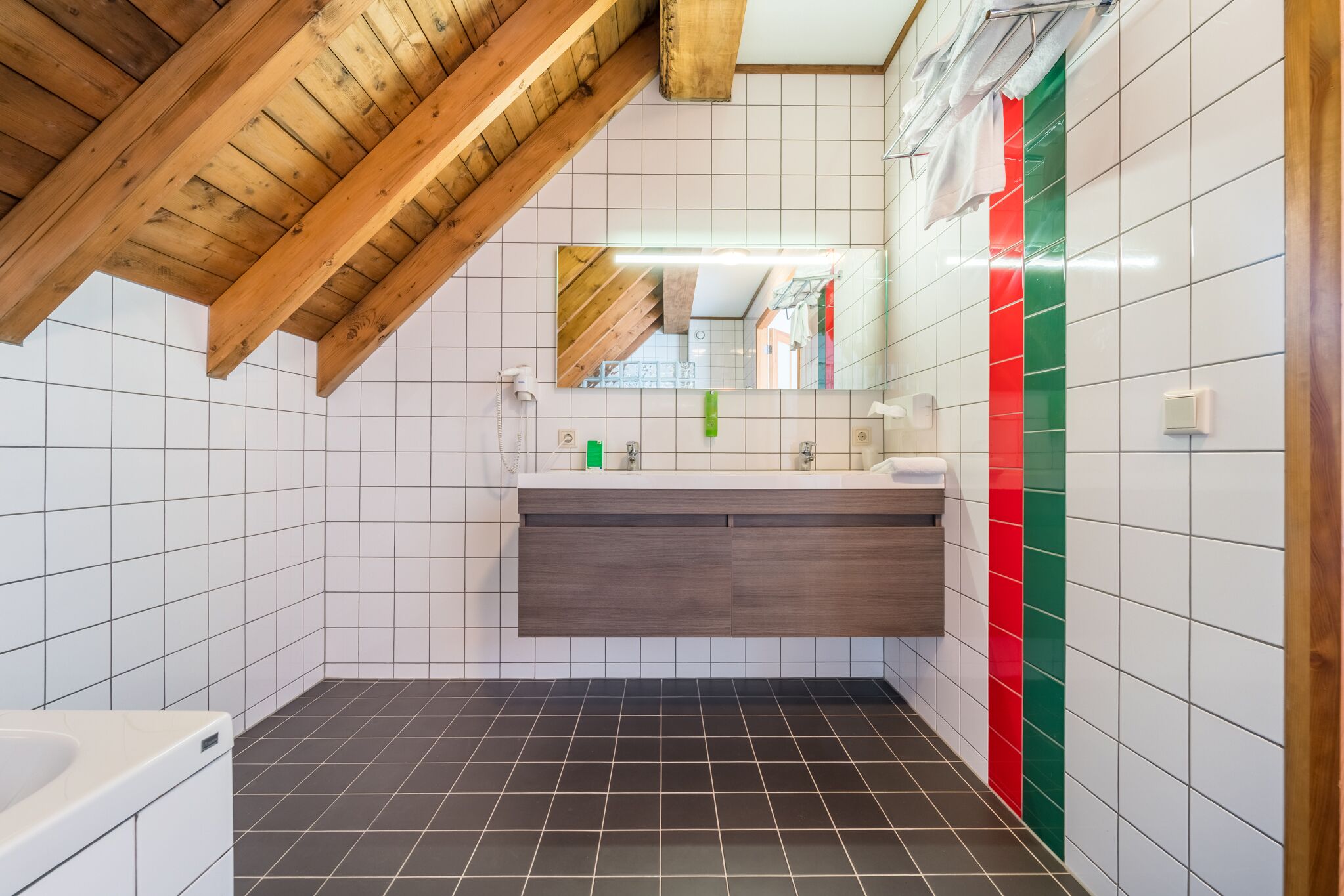 A modern bathroom with a wooden ceiling, white tiled walls, a dark tiled floor, a vanity with a mirror, and a red and green accent wall.