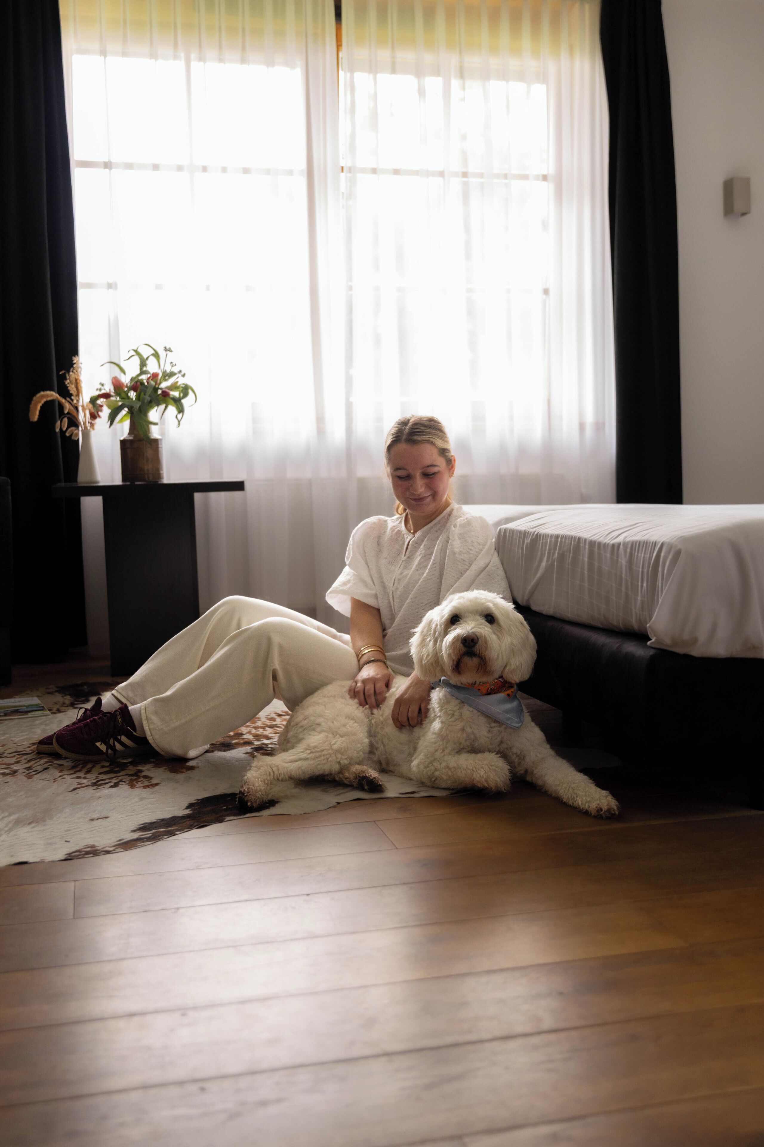 A person sitting on a rug next to a bed in a room with a wooden floor and a window with sheer curtains.