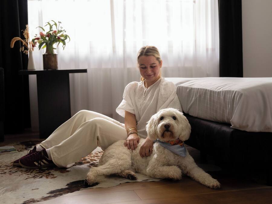 Una persona sentada sobre una alfombra junto a una cama en una habitación con piso de madera y una ventana con cortinas traslúcidas.