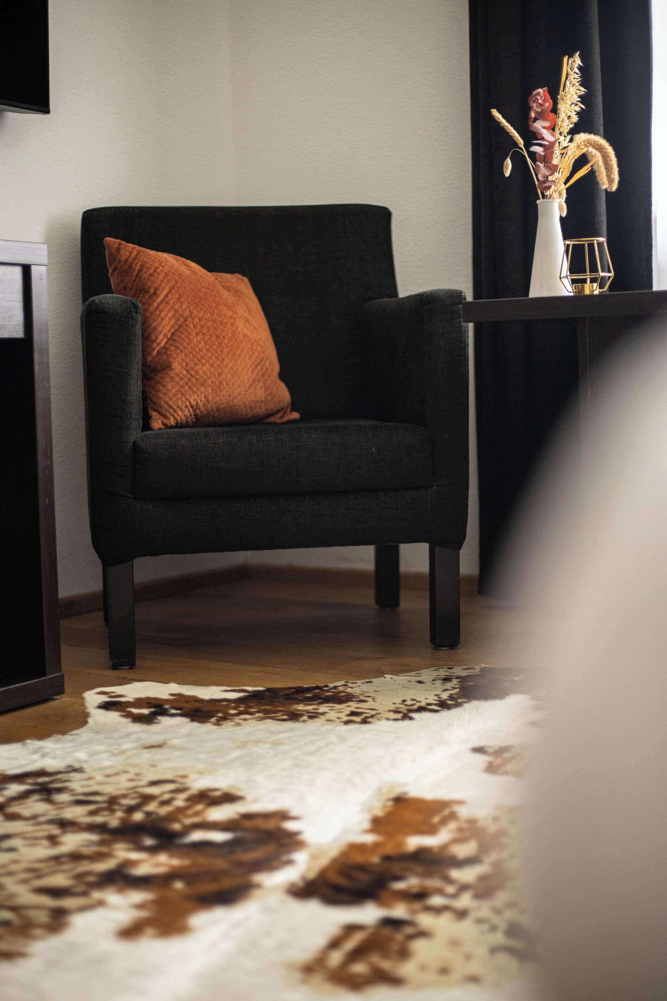 Una sala de estar moderna con un sillón oscuro, un tapete de cuero de vaca y un jarrón con plantas secas.