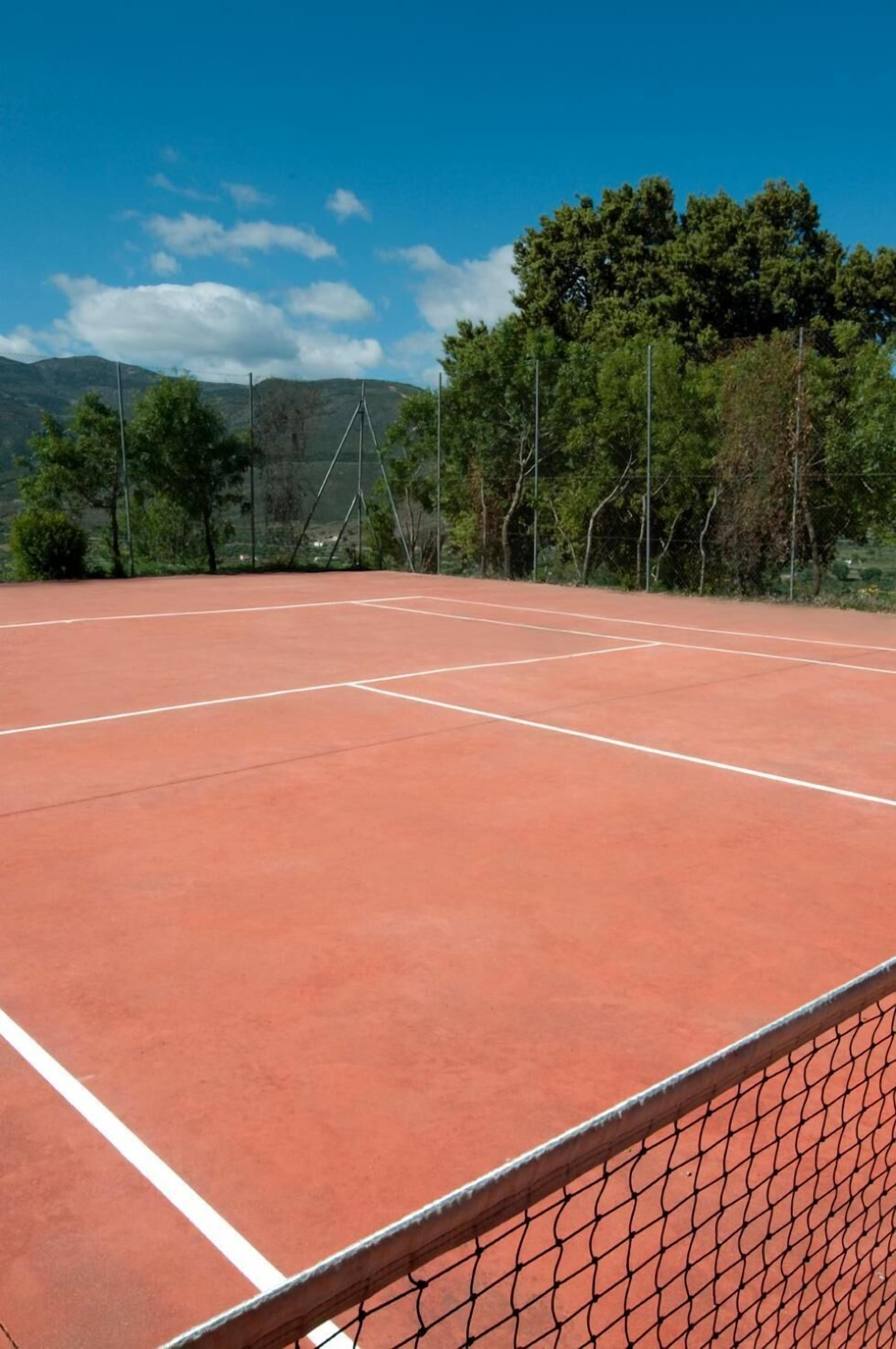 Una cancha de tenis con una red, rodeada de árboles y montañas al fondo.
