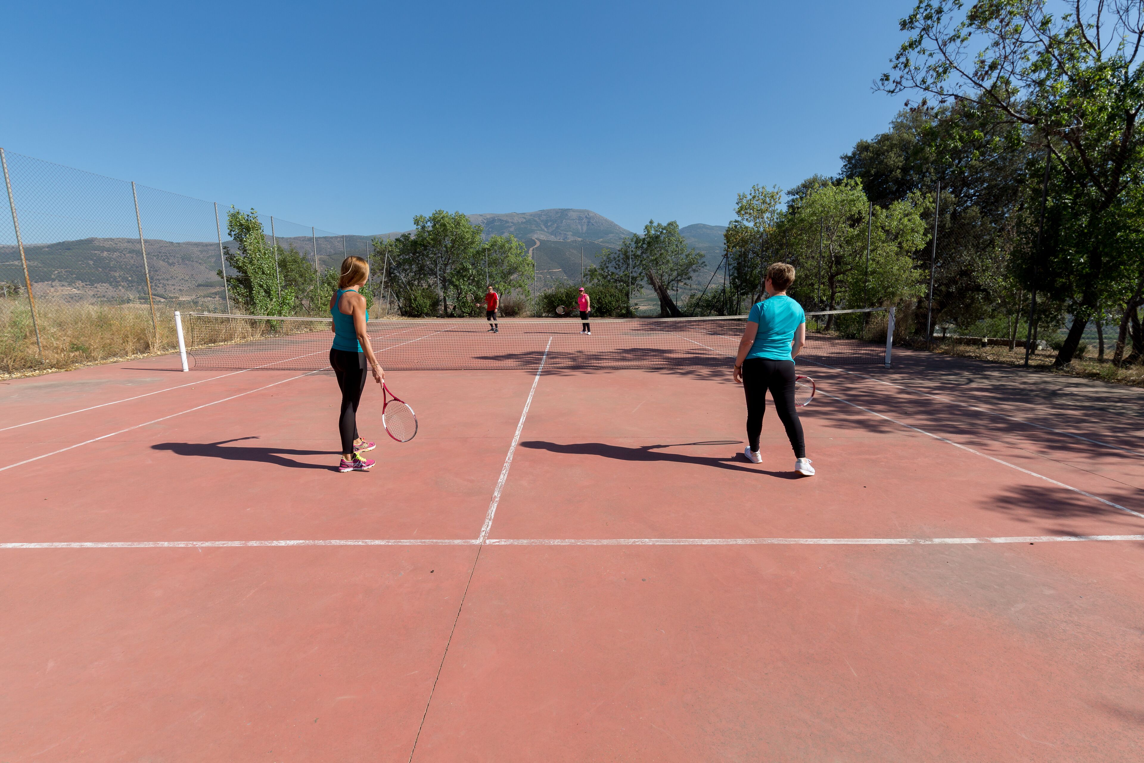 Un court de tennis avec un filet au milieu, entouré d’arbres et avec des montagnes en arrière-plan.