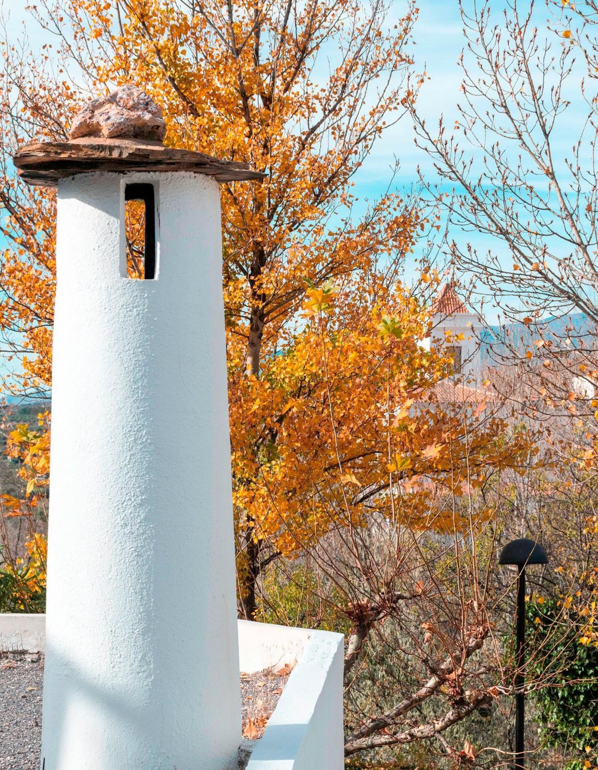 A white tower with a stone cap and a small window, surrounded by autumn-colored trees.