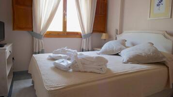 Una cama bien hecha con sábanas y almohadas blancas, una mesita de noche con lámpara y una ventana con cortinas.