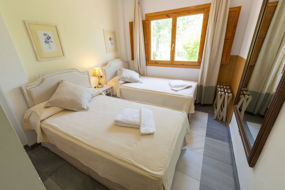A bedroom with two beds, a window with curtains, a bedside table with a lamp, and framed pictures on the wall.