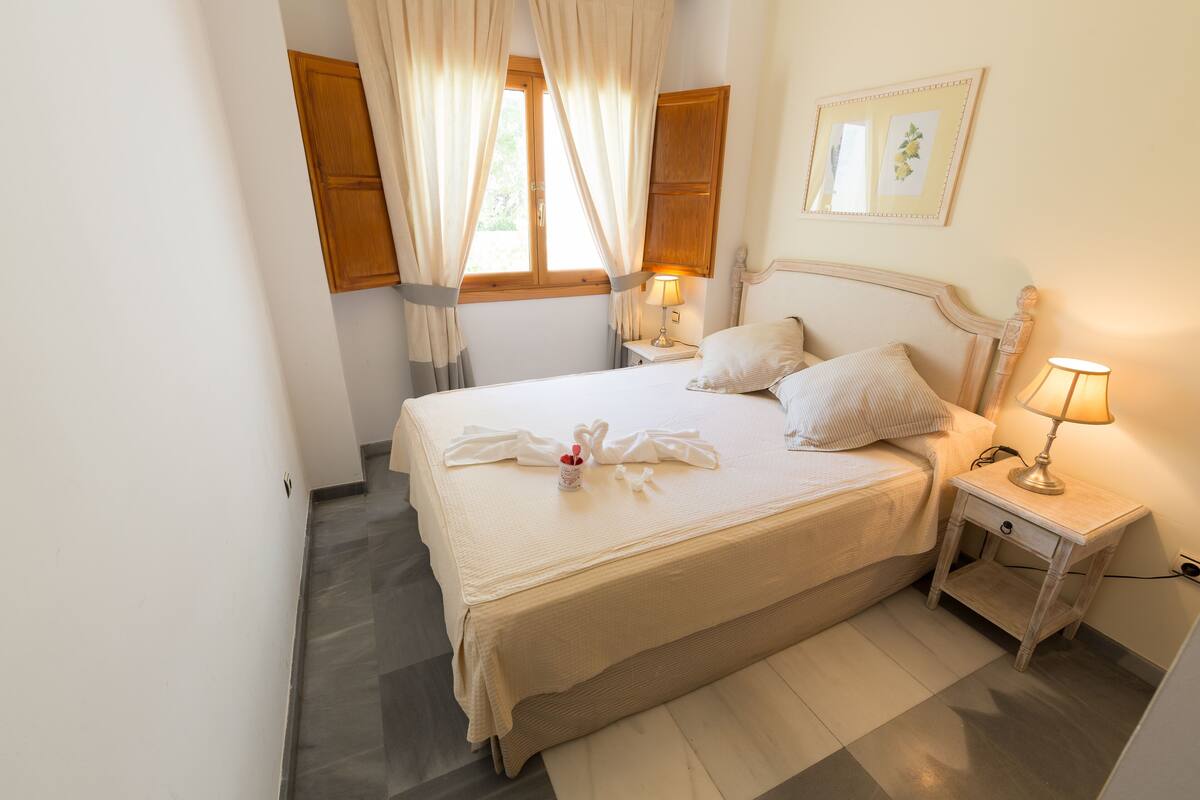 A neatly made bed with white linens and a towel arrangement, a bedside table with a lamp, and a window with curtains.
