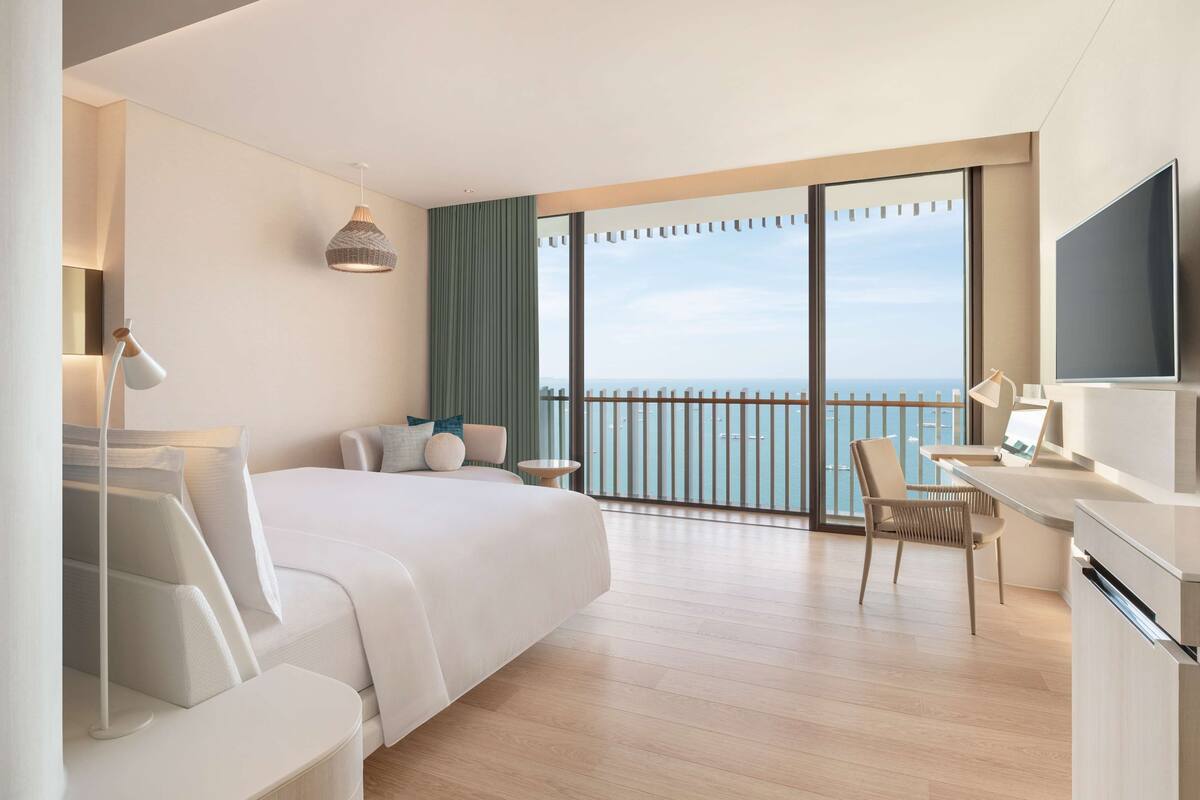 Ett modernt hotellrum med en stor säng, ett skrivbord, en stol, en TV och en balkong med utsikt över havet.