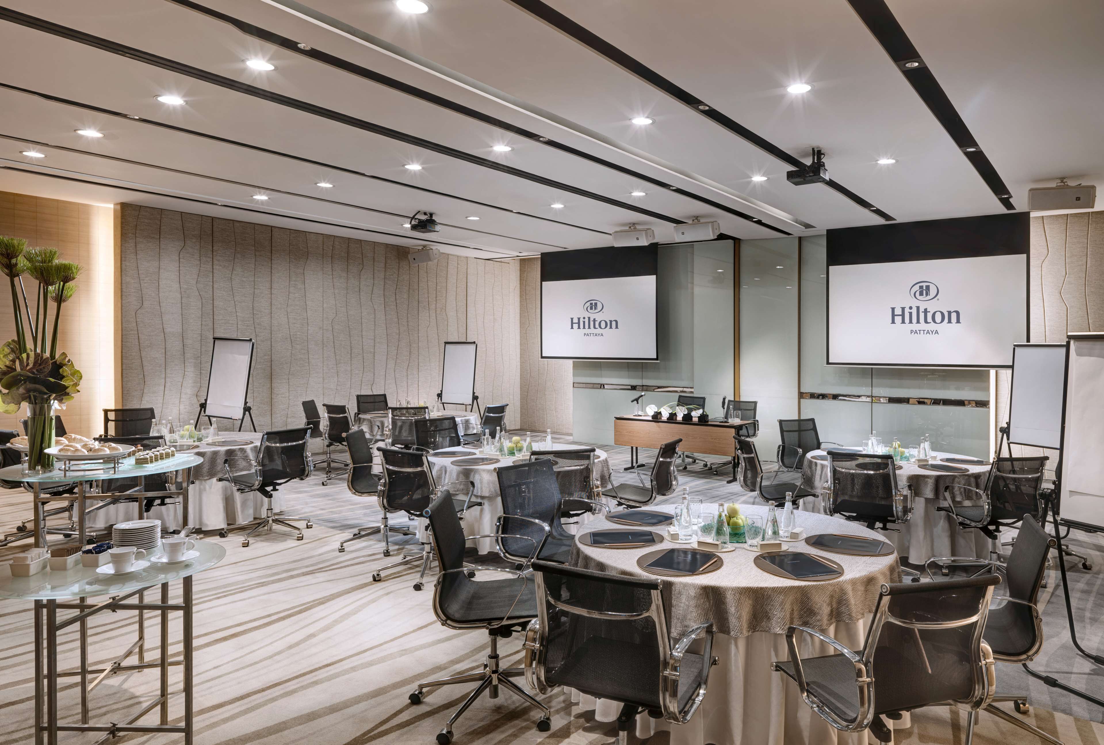 A conference room with multiple tables, chairs, and presentation screens displaying the Hilton logo.