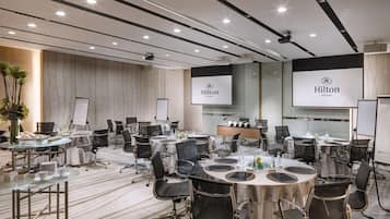 A conference room with multiple tables, chairs, and presentation screens displaying the Hilton logo.
