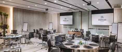 A conference room with multiple tables, chairs, and presentation screens displaying the Hilton logo.
