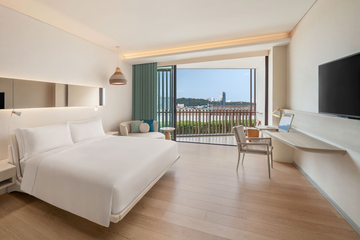 A modern hotel room with a large bed, a desk, a chair, a TV, and a balcony with a view of the city.