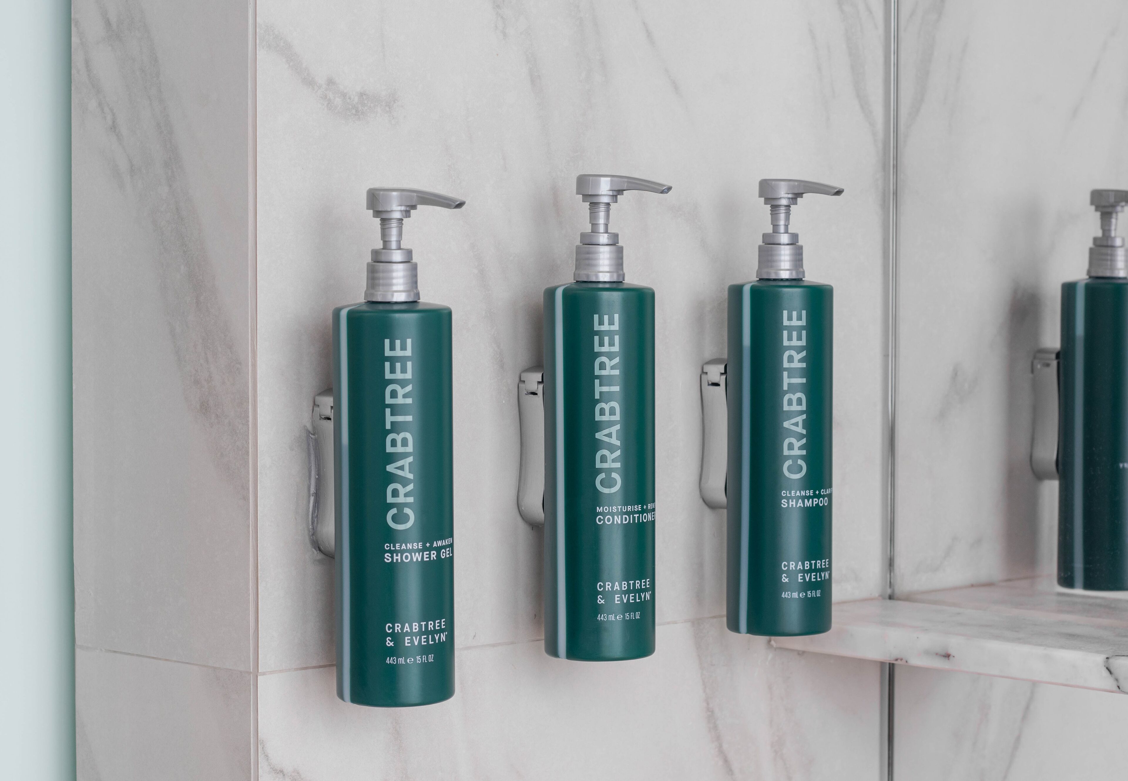 Three bottles of Crabtree shower gel and conditioner on a marble wall.
