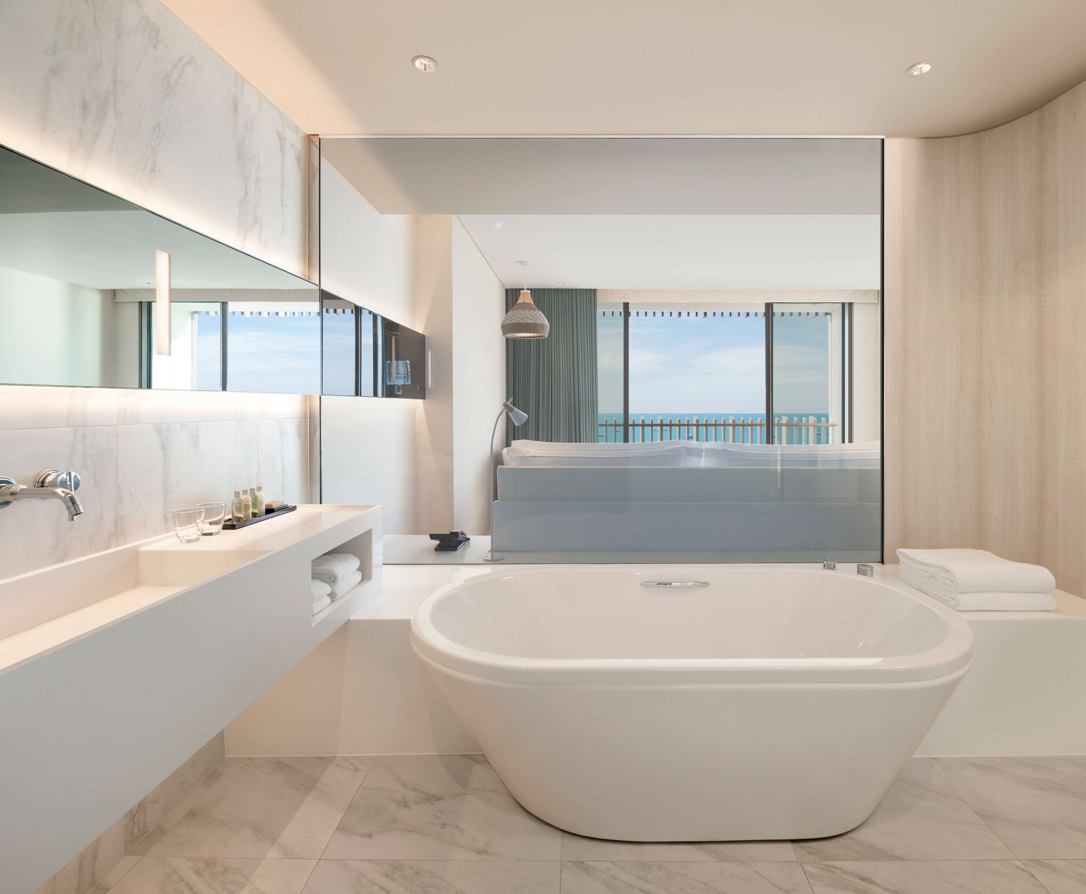 Un baño moderno con una bañera grande, un tocador con espejo y vistas al mar a través de un amplio ventanal.