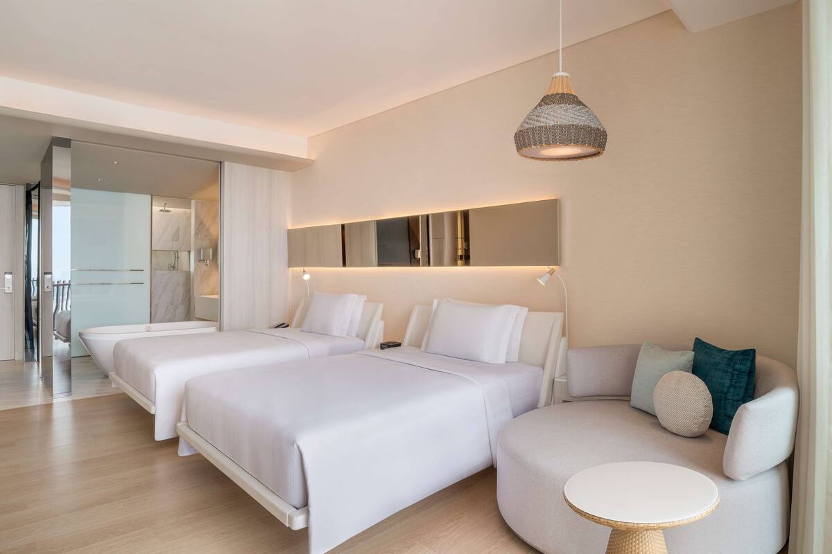 Ett modernt hotellrum med två sängar, en soffa, ett litet runt bord och en hängande lampa.