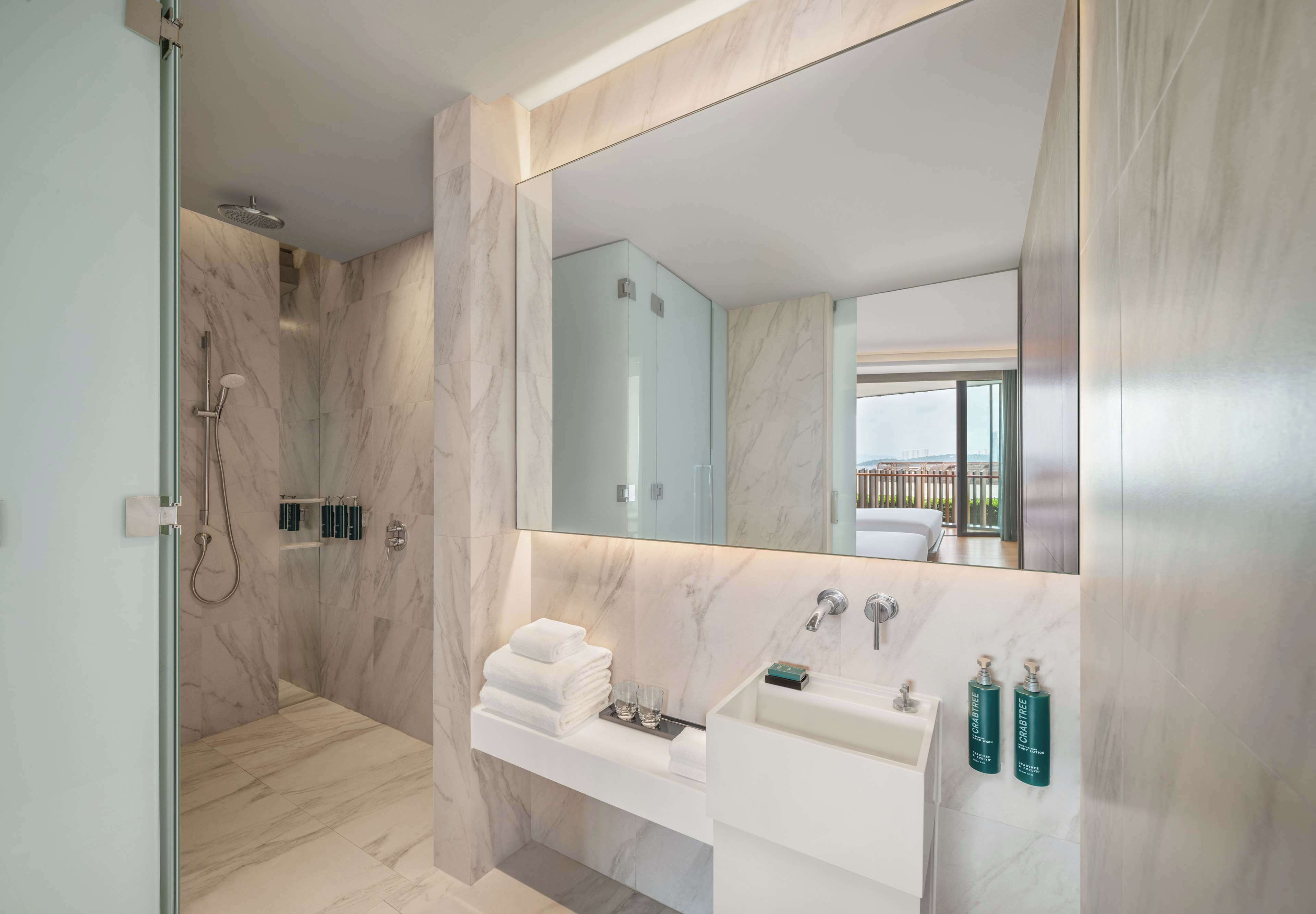 Un baño moderno con ducha de vidrio, paredes de mármol, un espejo grande, un lavamanos blanco con dos botellas de jabón para manos y vistas al exterior a través del espejo.