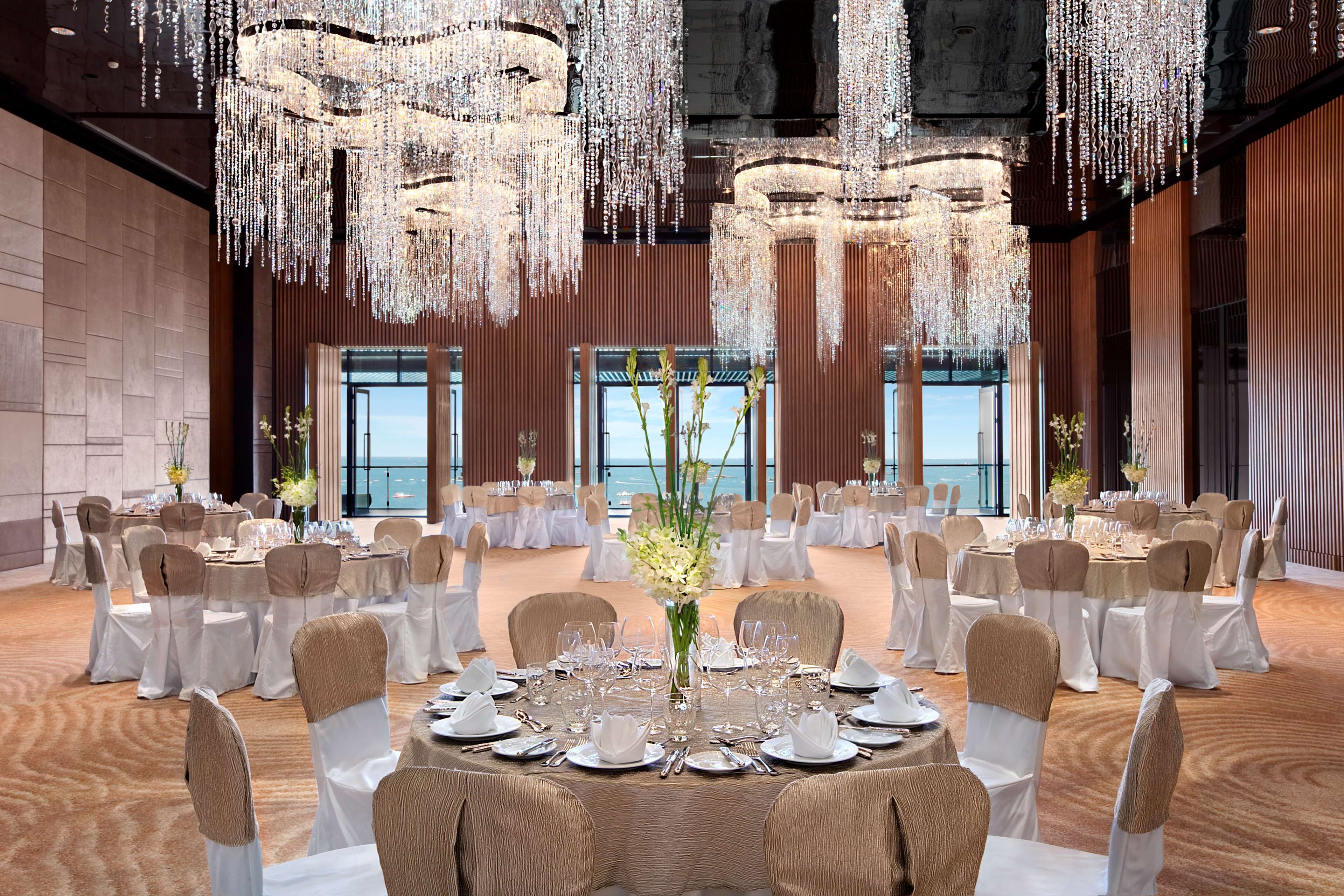 A banquet hall with round tables set for a formal event, featuring chandeliers and large windows overlooking the sea.