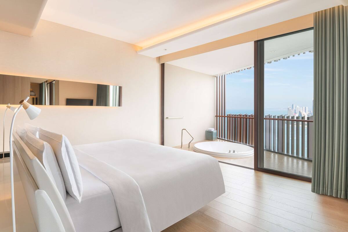 Ett modernt hotellrum med en stor säng, en sittgrupp med en fåtölj, ett litet bord, ett stort fönster med utsikt över staden och en balkong med ett räcke.
