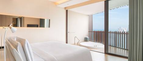 Una habitación de hotel moderna con una cama grande, una zona de estar con una silla, una mesita y un amplio ventanal con vistas a la ciudad, además de un balcón con barandilla.