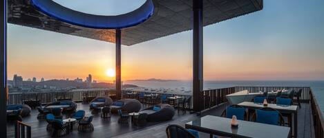 A rooftop bar with seating and a view of the sunset over the ocean.