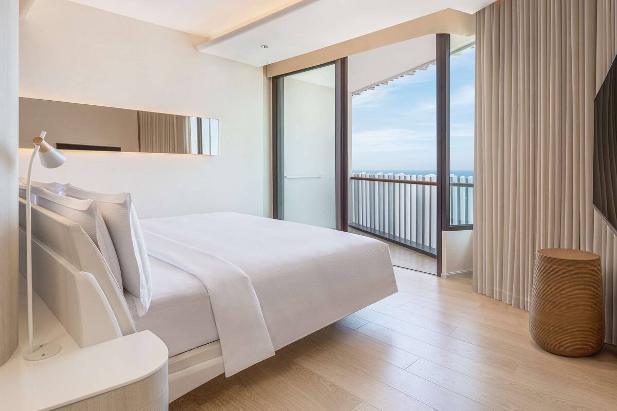 Ett modernt hotellrum med en stor säng, ett sängbord, en lampa, ett stort fönster med utsikt över himlen och en balkong med ett räcke.