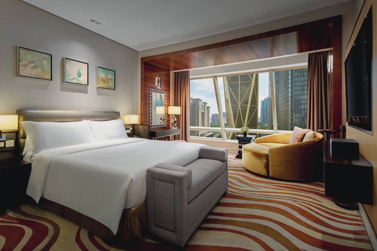 A hotel room with a large bed, a sofa, a chair, a large window with a city view, and artwork on the wall.