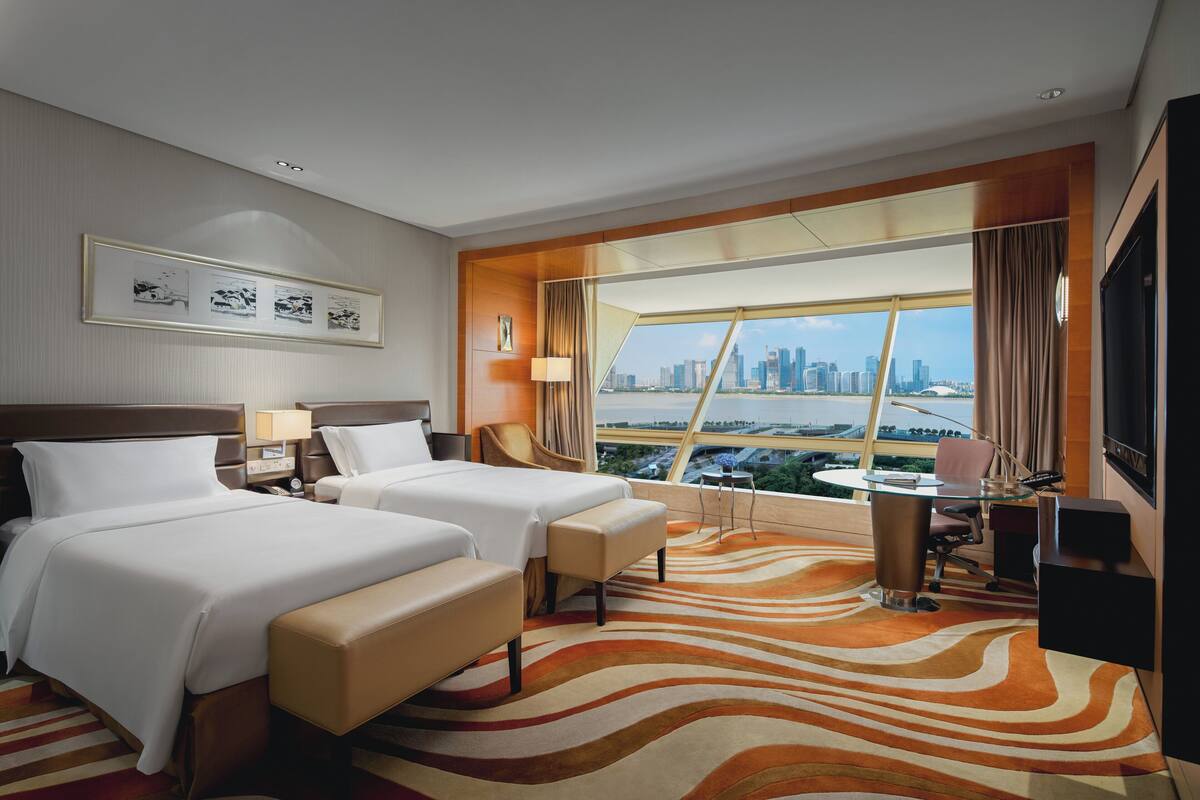A hotel room with two beds, a desk, and a view of a cityscape.