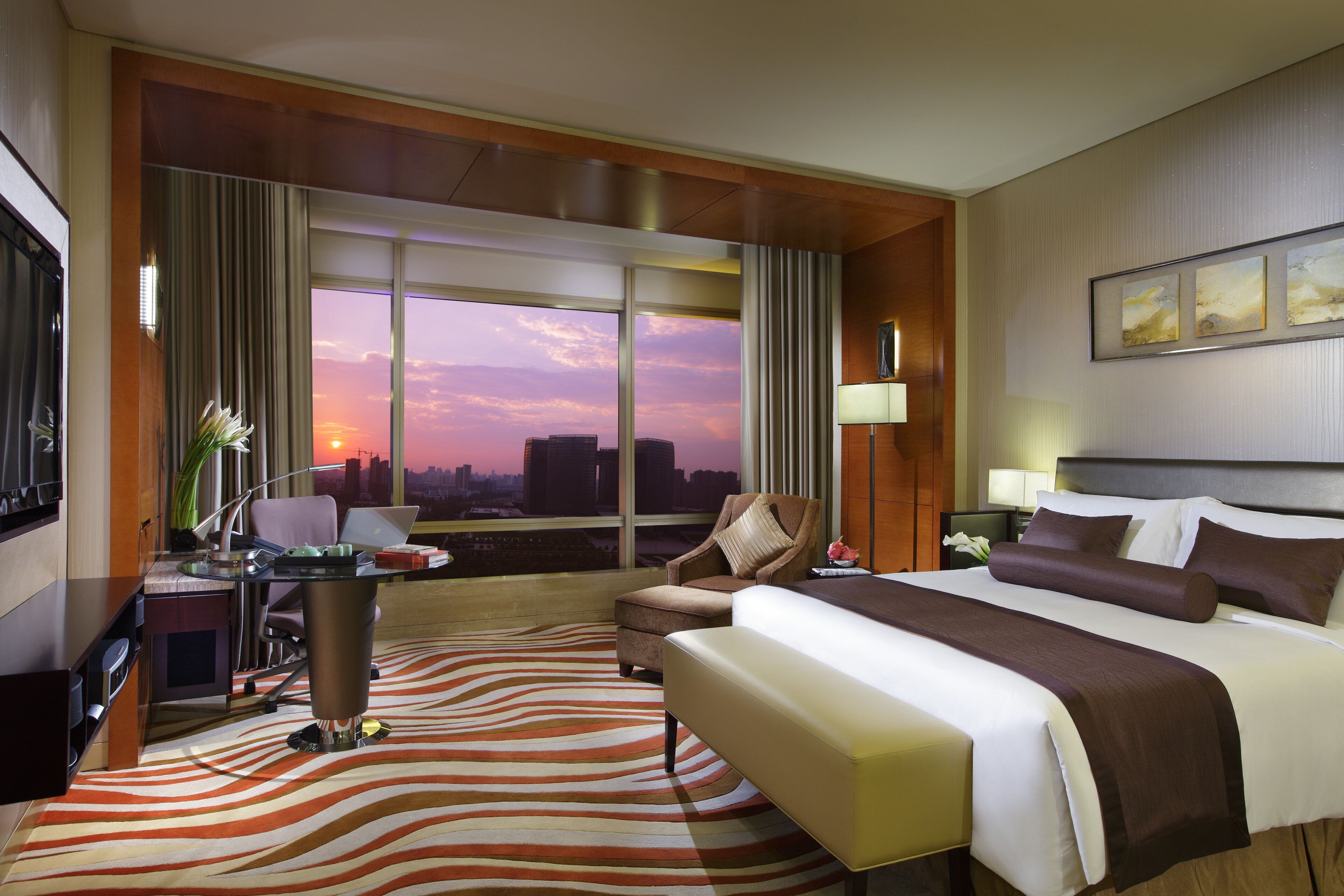 Ein Hotelzimmer mit Bett, Schreibtisch, Stuhl, Fernseher und einem großen Fenster mit Blick auf die Stadt.